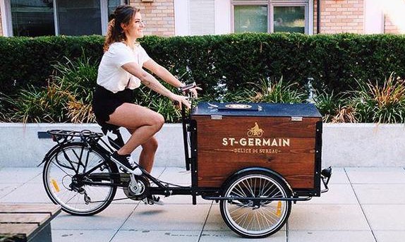 A Peddler pedaling a St-germain branded custom cedar cargo box delivery bike down the sidewalk on a sunnuy day 