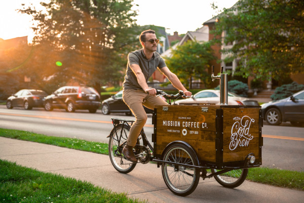 A man riding a cold brew coffee bike / trike down the side walk 