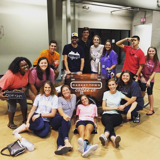 A group of smiling people pose with a coffee cart bike in a gym