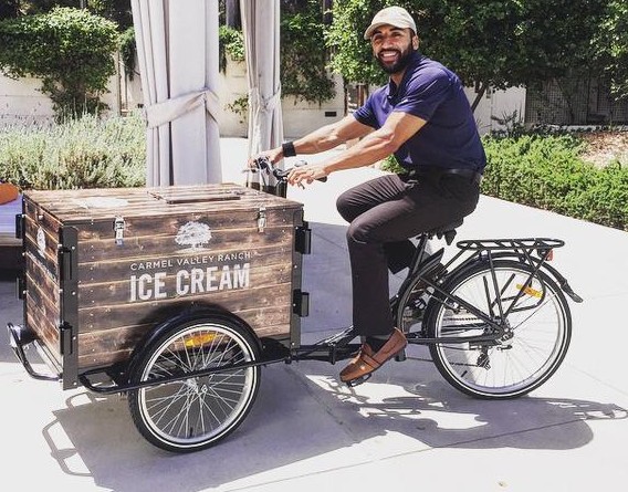 A man riding a Carmel Valley Ranch branded Ice Cream trike / bike down the sidewalk.