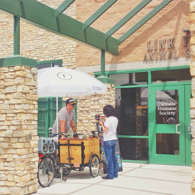 A barista is serving a customer from a custom cedar box coffee bike parked in front of the humane society