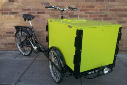 an Icicle Tricycles Custom Cargo Bike with the panels wrapped with green vinyl wrap parked on the sidewalk in front of a brick wall