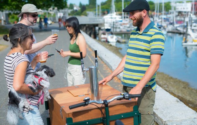 A man smiling at a woman holding a dog standing above a cedar wood cargo box cold brew coffee bike / trike at a marina.