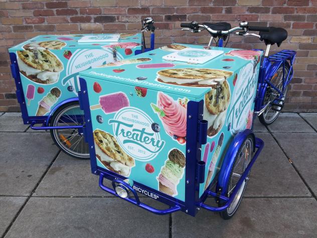 Two Icicle Tricycles Ice Cream Tricycles / Bicycles branded The Treatery parked on the sidewalk in front of a brick wall