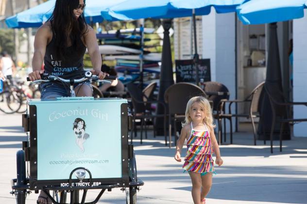 a woman riding an Icicle Tricycles Marketing Ice Cream Bicycle / tricycle branded for Coconut Girl Paleo Ice Cream down the side walk next to a toddler