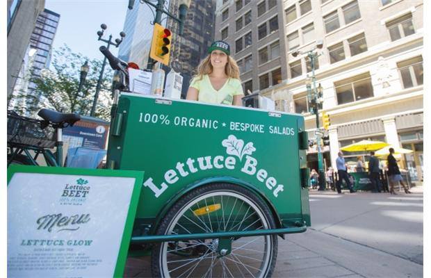 A woman standing behind icicle tricycles front load food bike / cart branded for the lettuce beet / bike salad bike at a popup event in the city