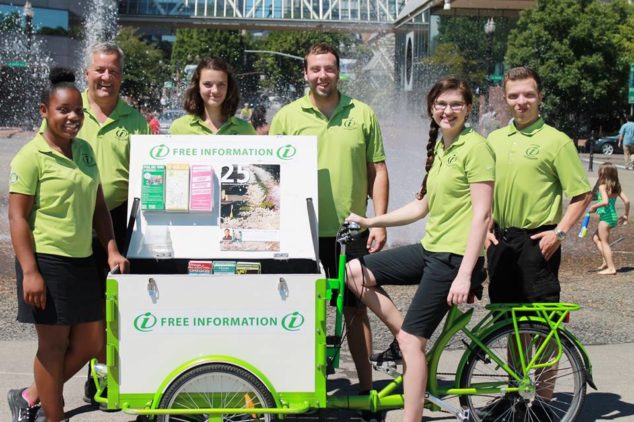 Smiling team of people with mobile information kiosk ice cream bike in front of a fountain.