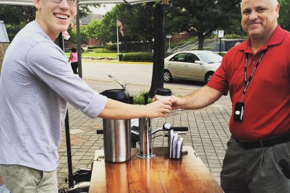 A Smiling man serving hot coffee from a wood wrapped cold brew coffee bike / trike 