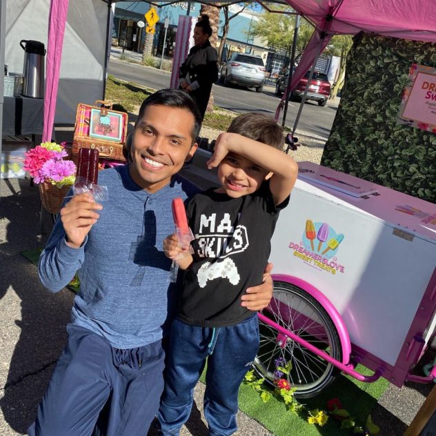 father and son smiling with popsicles with ice cream in front of an ice cream cart at a farmers market 