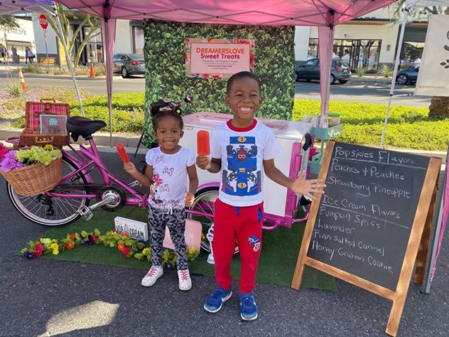 two Children smiling with popsicles with ice cream in front of an ice cream cart at a farmers market 