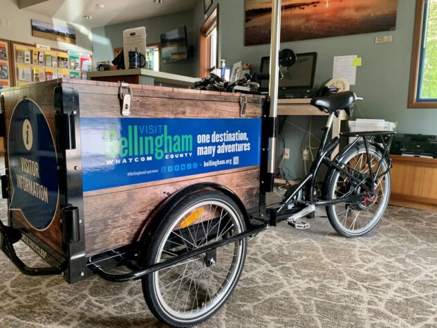 A Mobile Tourist Information Bike for Bellingham, Washington sits in a carpeted room. Behind it are a desk and counter with office supplies on them.
