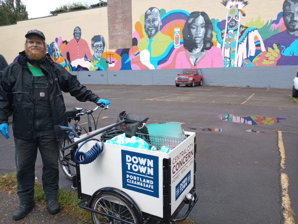 A Portland Clean & Safe worker proudly stands next their mobile street cleaning bike. The cargo area of the bike is filled with cleaning equipment.