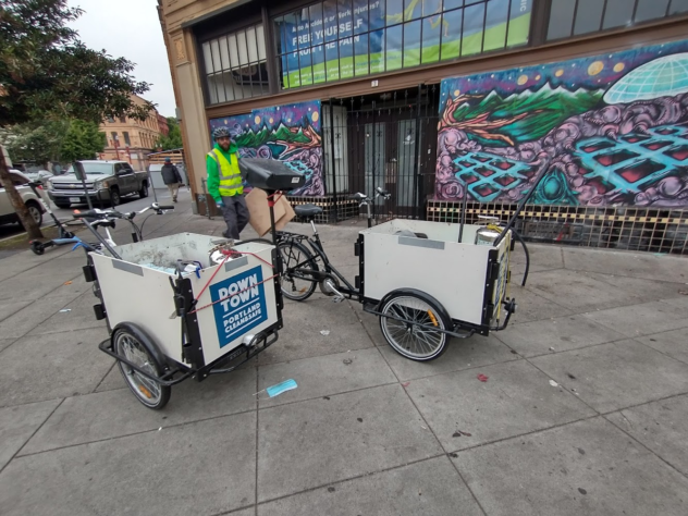 A Portland Clean & Safe worker walks behind two mobile street cleaning bikes, carrying trash from a cleanup.