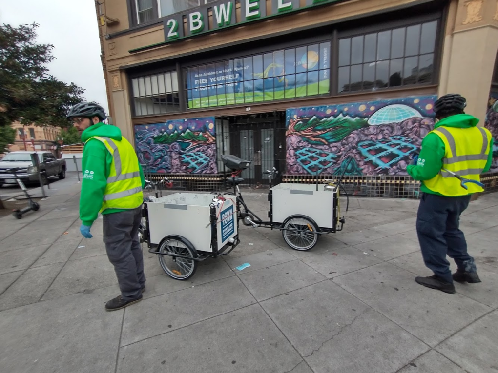 Two Clean & Safe workers picking up trash in front of two Mobile Graffiti Removal Bikes. There is a graffiti mural in the background.