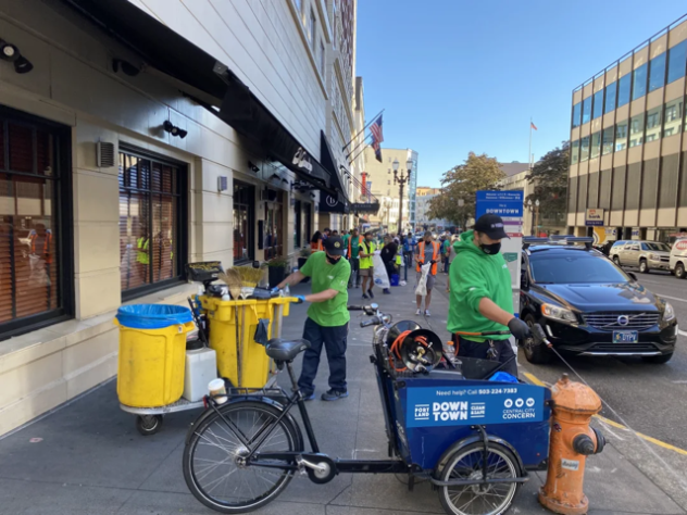 A member of a city Graffiti Removal team maneuvers a handcart with street cleaning supplies and trashcans along a crowded sidewalk while another team member sprays water from the pressure washer on their Graffiti Removal Bike.